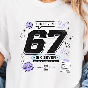 Pu&ograve; includere: Una t-shirt bianca a maniche corte con una grande grafica nera del numero 67. Il design include accenti viola e neri, il testo SIX SEVEN, una faccina sorridente, una corona, una clessidra e una nota con scritto 67% CHAOS 33% CONFIDENCE.