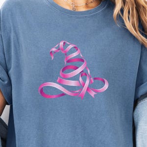 May include: A blue jean colored t-shirt featuring a graphic of a witch hat constructed from a pink ribbon. The shirt has a crew neckline and short sleeves. A circular badge in the upper right corner contains the text blue jean.