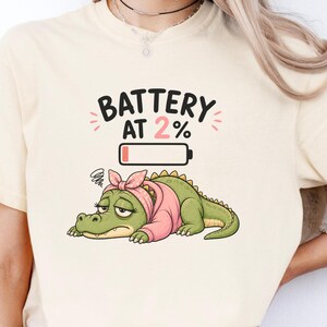 May include: An ivory short-sleeve t-shirt featuring a graphic of a tired green alligator wearing a pink headband. Above the alligator is a battery icon and the text "BATTERY AT 2%" in black lettering. The shirt has a crew neckline and a relaxed fit.