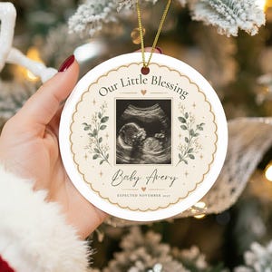 Puede incluir: Un adorno redondo de cerámica blanca con un borde dorado festoneado, una imagen de ecografía en blanco y negro en el centro y el texto Our Little Blessing, Baby Avery y EXPECTED NOVEMBER 2027, decorado con ilustraciones botánicas verdes y estrellas doradas.