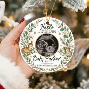 Puede incluir: Un adorno redondo de cerámica blanca con una ecografía en el centro, rodeado por una corona botánica verde con un ciervo y un conejo. El texto dice: Hello Little One, Baby Parker, Expected Arrival, See You Soon, 2027. Cordón dorado incluido.