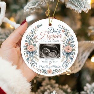 May include: A round white ceramic ornament featuring a central ultrasound image, surrounded by a floral wreath of pink flowers and blue leaves. Text reads: Baby Harper COMING SOON, EXPECTED APRIL 18, 2027, and Our Tiny Miracle. Gold string attached.
