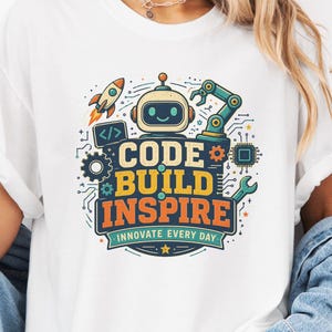 May include: A white t-shirt featuring a colorful graphic print of a smiling robot, a rocket, and mechanical parts. The text reads CODE BUILD INSPIRE INNOVATE EVERY DAY in bold, multi-colored lettering. The design includes circuit board patterns.