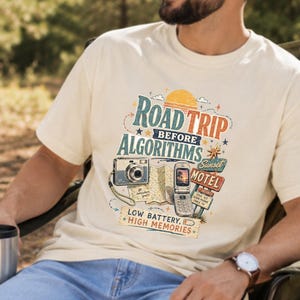 May include: An ivory short-sleeve t-shirt featuring a graphic print with the text ROAD TRIP BEFORE ALGORITHMS, SUNSET MOTEL, and LOW BATTERY, HIGH MEMORIES. The design includes illustrations of a vintage camera, a map, and a flip phone.