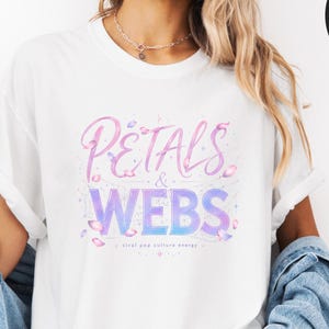 May include: A white short-sleeved t-shirt featuring the text Petals & Webs in a gradient pink and purple font. The design includes small petal illustrations and sparkles. The shirt is worn with a blue denim jacket and denim shorts.
