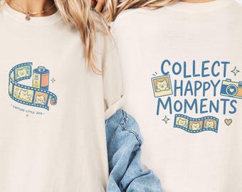 May include: An ivory t-shirt featuring a small graphic on the front of a film roll with cat illustrations and the text "CAPTURE LITTLE JOYS". The back displays a larger graphic with the text "COLLECT HAPPY MOMENTS", a camera, a photo, and film strips.