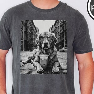 Peut inclure: Un t-shirt &agrave; col rond gris anthracite avec une impression graphique en noir et blanc repr&eacute;sentant un chien portant des lunettes de soleil et buvant dans un gobelet en plastique avec une paille, sur fond de rue urbaine. Le mot pepper est inscrit.