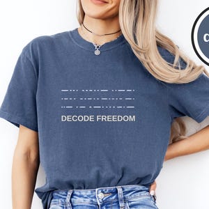 May include: Denim blue t-shirt with the text "DECODE FREEDOM" in white. The shirt features a Morse code design above the text. The shirt is paired with blue jeans. A denim-colored circle with the word "denim" is in the upper right corner.