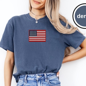 Può includere: Una t-shirt a maniche corte con girocollo in un colore blu denim sbiadito. Al centro del petto è presente una toppa rettangolare ricamata della bandiera degli Stati Uniti nei colori rosso, bianco e blu. Un badge circolare mostra la parola denim.
