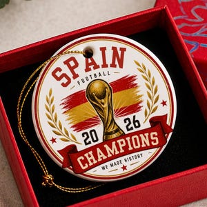 Könnte beinhalten: Ein rundes, weißes Keramikornament mit goldener Aufhängeschnur, verziert mit einer goldenen Trophäe, der spanischen Flagge und dem Text SPAIN FOOTBALL 2026 CHAMPIONS WE MADE HISTORY. Das Ornament liegt in einer roten Geschenkbox.