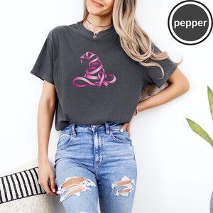May include: A charcoal gray short-sleeve t-shirt featuring a graphic of a witch hat constructed from a pink awareness ribbon. The shirt has a crew neckline and a relaxed fit. A circular logo in the top right corner contains the text pepper.