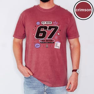 Pu&ograve; includere: Una t-shirt a maniche corte color cremisi con una grande grafica nera del numero 67 e il testo SIX SEVEN, NOT JUST A NUMBER. IT'S A MOOD. Le grafiche aggiuntive includono una faccina sorridente, una corona, stelle, una clessidra e un foglietto.