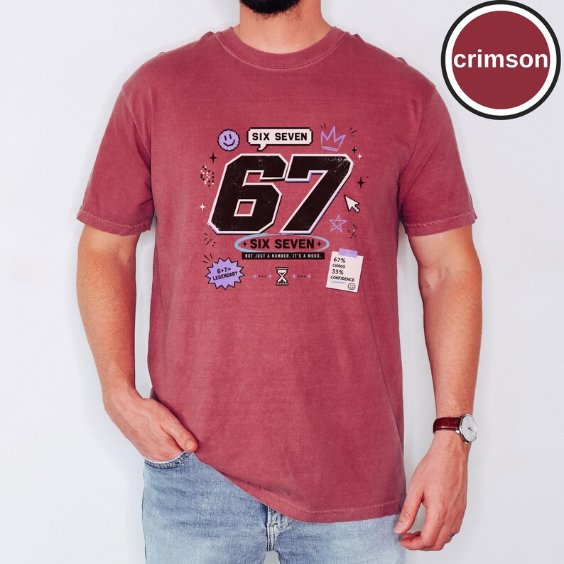 Pu&ograve; includere: Una t-shirt a maniche corte color cremisi con una grande grafica nera del numero 67 e il testo SIX SEVEN, NOT JUST A NUMBER. IT'S A MOOD. Le grafiche aggiuntive includono una faccina sorridente, una corona, stelle, una clessidra e un foglietto.