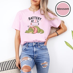 May include: A light pink t-shirt featuring a graphic of a tired green crocodile wearing a pink headband and sweater. Above the crocodile, the text reads BATTERY AT 2% with a low battery icon. A circular logo in the top right corner says blossom.