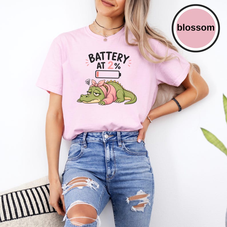 May include: A light pink t-shirt featuring a graphic of a tired green crocodile wearing a pink headband and sweater. Above the crocodile, the text reads BATTERY AT 2% with a low battery icon. A circular logo in the top right corner says blossom.