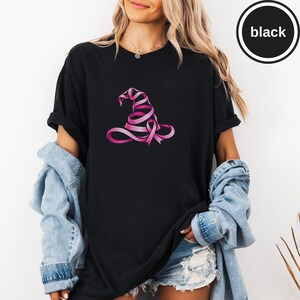 May include: A black short-sleeved t-shirt featuring a graphic of a witch hat constructed from a pink awareness ribbon. The shirt has a crew neckline and a relaxed fit. A circular badge in the upper right corner contains the text black.