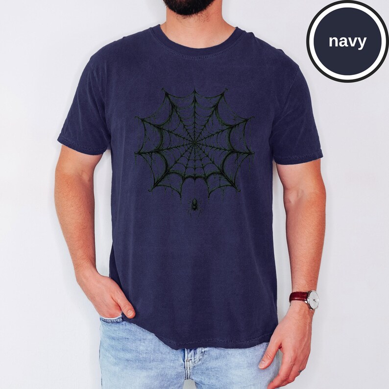 May include: A navy blue short-sleeved t-shirt featuring a black graphic print of a detailed spider web centered on the chest. Below the web, a small black spider is depicted. The shirt has a crew neckline and a standard fit, with the word navy visible.