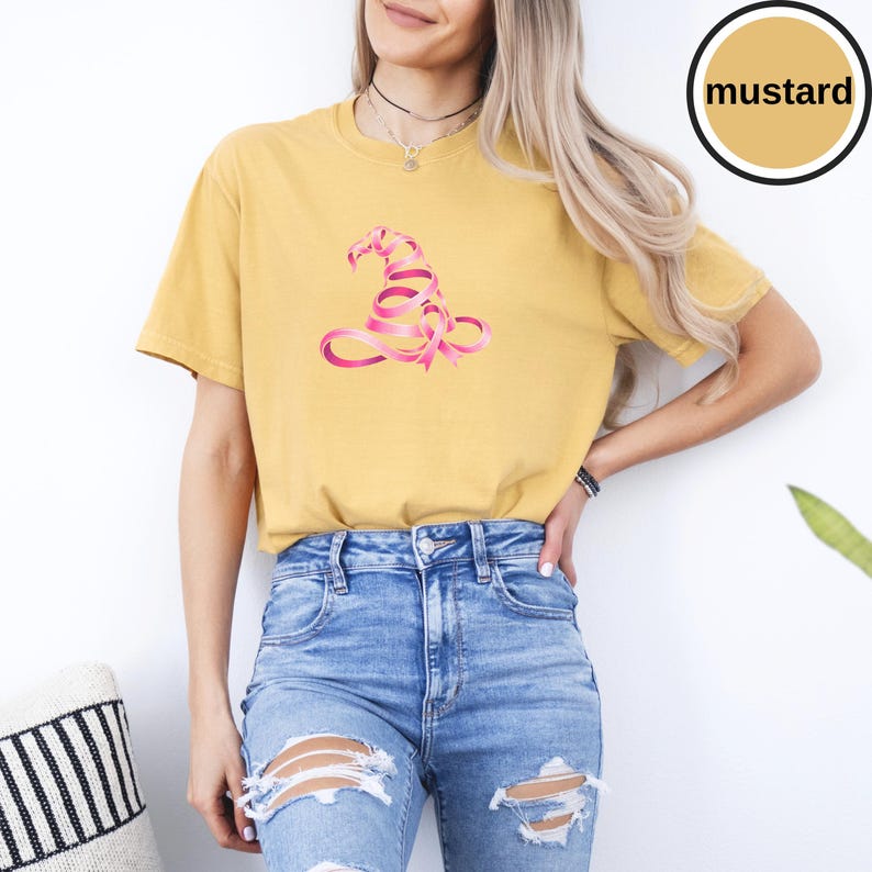 May include: A mustard yellow short-sleeve t-shirt featuring a graphic of a witch hat formed from a pink ribbon. The shirt has a crew neckline and a relaxed fit. A circular badge in the upper right corner contains the text mustard.