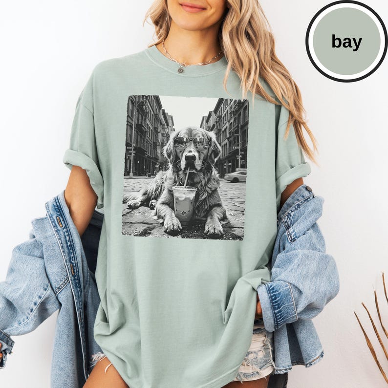 Peut inclure: Un t-shirt vert sauge avec une impression graphique en noir et blanc repr&eacute;sentant un chien portant des lunettes de soleil et buvant dans un gobelet en plastique avec une paille, sur fond de rue urbaine. Le t-shirt a un col rond.