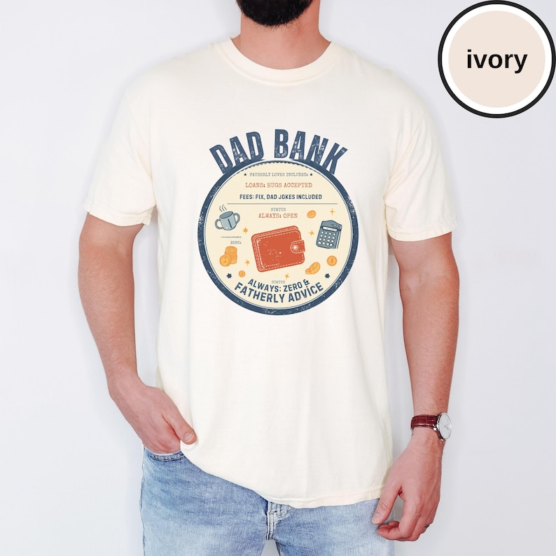 May include: Ivory-colored t-shirt with a circular graphic that reads "DAD BANK." The graphic includes a wallet, calculator, coins, and a coffee cup. The text also says "Loans Hugs Accepted" and "Always Zero & Fatherly Advice."