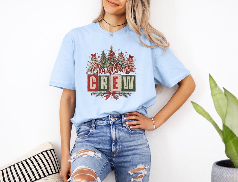 Op de afbeelding: Een lichtblauw t-shirt met korte mouwen en een grafische print van verschillende gestileerde kerstbomen boven de tekst "Christmas CREW" in rode en groene blokletters, geaccentueerd met een rode strik onderaan het ontwerp.