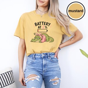 May include: A mustard yellow short-sleeve t-shirt featuring a graphic of a tired green alligator wearing a pink headscarf. Above the alligator is a low battery icon and the text BATTERY AT 2%. The shirt has a crew neckline and a relaxed fit.