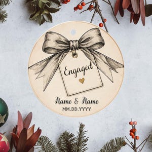 May include: A round, cream-colored ornament featuring a black and white sketch of a large bow tied to a gift tag. The tag displays the word Engaged in black script with a small gold heart below it, followed by the text Name & Name and MM.DD.YYYY.