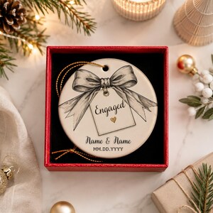May include: A round white ceramic ornament inside a red gift box. The ornament features a black line-art illustration of a bow tied to a gift tag with the word Engaged and a small gold heart. Below, the text reads Name & Name and MM.DD.YYYY.