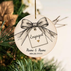 May include: A round ceramic ornament featuring a black and white sketch of a large bow tied to a gift tag. The tag displays the word Engaged with a small gold heart. Below, the text Name & Name and MM.DD.YYYY is printed in black ink.