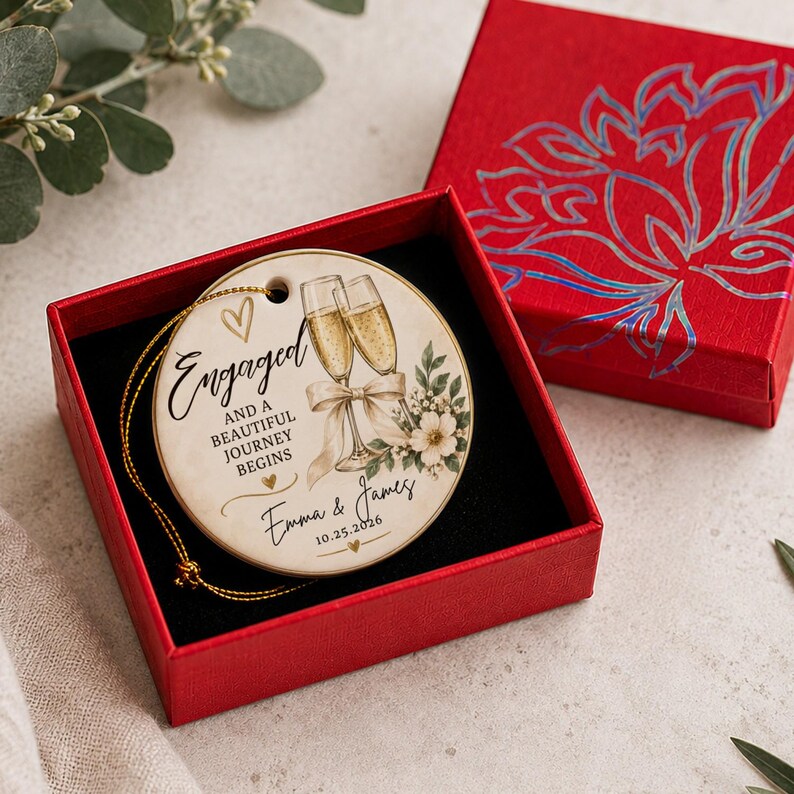K&ouml;nnte beinhalten: Ein runder Keramikschmuck mit goldener Schnur in einer roten Geschenkbox. Der Schmuck zeigt zwei Champagnergl&auml;ser, eine Schleife, florale Akzente und den Text: Engaged and a beautiful journey begins, Emma & James, 25.10.2026, mit kleinen Herzsymbolen.