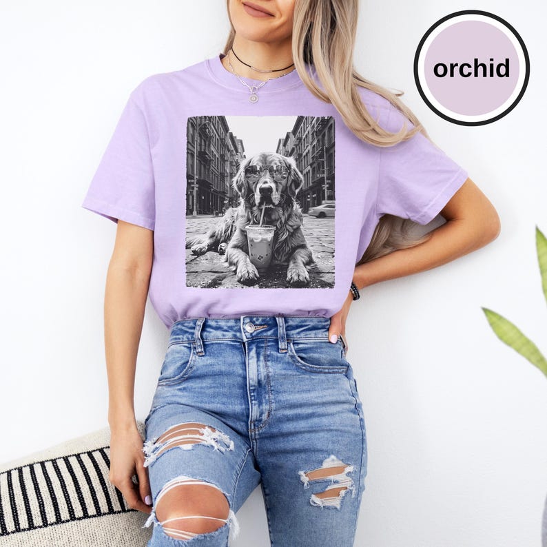Peut inclure: Un t-shirt &agrave; manches courtes violet clair avec une impression graphique en noir et blanc repr&eacute;sentant un chien portant des lunettes de soleil et buvant dans un gobelet sur une rue urbaine. Le t-shirt a un col rond. Le mot orchid est imprim&eacute;.