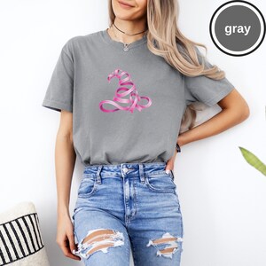 May include: A gray short-sleeved crew neck t-shirt featuring a graphic of a witch hat constructed from a pink ribbon. The shirt has a relaxed fit and is paired with blue denim jeans that have distressed rips at the knees and thighs.