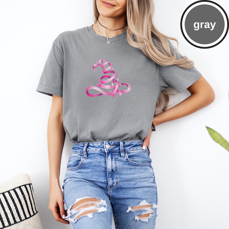 May include: A gray short-sleeved crew neck t-shirt featuring a graphic of a witch hat constructed from a pink ribbon. The shirt has a relaxed fit and is paired with blue denim jeans that have distressed rips at the knees and thighs.
