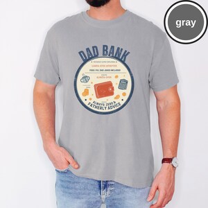May include: Gray t-shirt with a circular graphic that reads "DAD BANK." The graphic includes a wallet, calculator, and coffee cup, with text stating "Loans: Hugs Accepted" and "Always: Zero & Fatherly Advice."