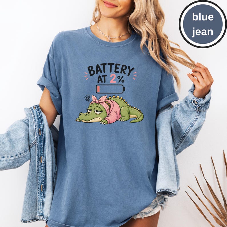May include: A blue jean coloured t-shirt featuring a graphic of a tired green crocodile wearing a pink headscarf and sweater. Above the crocodile, the text reads BATTERY AT 2% with a low battery icon. The shirt has a crew neckline and short sleeves.