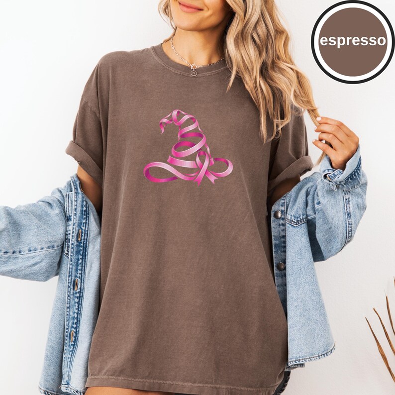 May include: An espresso-colored short-sleeve t-shirt featuring a graphic of a witch hat formed from a pink ribbon. The shirt is worn with a light blue denim jacket. A circular badge in the top right corner contains the text espresso.