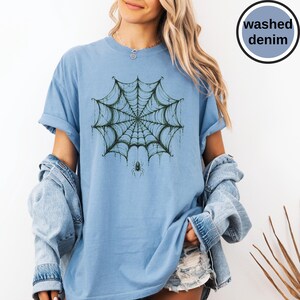 May include: A light blue t-shirt featuring a dark green spider web graphic with a small spider hanging from the center. The shirt has a crew neckline and short sleeves. A circular badge in the top right corner contains the text washed denim.