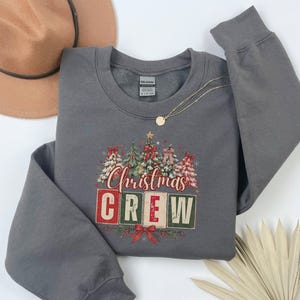 Op de afbeelding: Een donkergrijze sweater met ronde hals en een afbeelding van vijf gestileerde kerstbomen met rode strikken en de tekst "Christmas CREW" in rode en groene blokletters. Het ontwerp bevat een kleine gouden ster boven op de middelste boom.