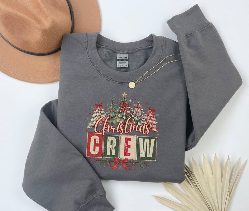 Op de afbeelding: Een donkergrijze sweater met ronde hals en een afbeelding van vijf gestileerde kerstbomen met rode strikken en de tekst "Christmas CREW" in rode en groene blokletters. Het ontwerp bevat een kleine gouden ster boven op de middelste boom.