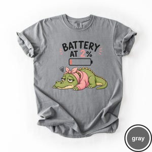 May include: A grey t-shirt featuring a graphic of a tired green crocodile wearing a pink headscarf. Above the crocodile is the text "BATTERY AT 2%" and a low battery icon with a small red bar, indicating a low charge level for the character.