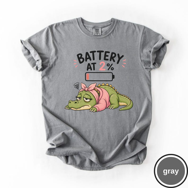 May include: A grey t-shirt featuring a graphic of a tired green crocodile wearing a pink headscarf. Above the crocodile is the text "BATTERY AT 2%" and a low battery icon with a small red bar, indicating a low charge level for the character.
