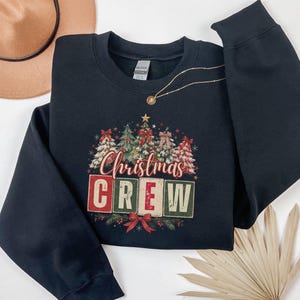 Op de afbeelding: Een zwart sweatshirt met lange mouwen en ronde hals met een grafische print van verschillende versierde kerstbomen boven de tekst "Christmas CREW" in rode, groene en cr&egrave;mekleurige blokletters, geaccentueerd met een rode strik onderaan.