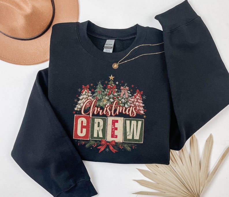 Op de afbeelding: Een zwart sweatshirt met lange mouwen en ronde hals met een grafische print van verschillende versierde kerstbomen boven de tekst "Christmas CREW" in rode, groene en cr&egrave;mekleurige blokletters, geaccentueerd met een rode strik onderaan.