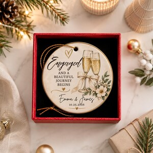 K&ouml;nnte beinhalten: Ein rundes, wei&szlig;es Keramikornament mit goldener Schnur, bedruckt mit zwei Champagnergl&auml;sern, einer Schleife und Blumenmotiven. Der Text lautet: Engaged and a beautiful journey begins, Emma & James, 10.25.2026. Es liegt in einer roten Schachtel.
