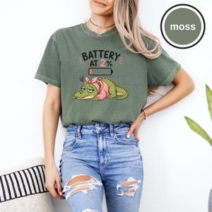 May include: A moss green short-sleeve t-shirt featuring a graphic of a tired green alligator wearing a pink headband. Above the alligator, the text reads BATTERY AT 2% with a low battery icon. The shirt has a crew neckline and a relaxed fit.