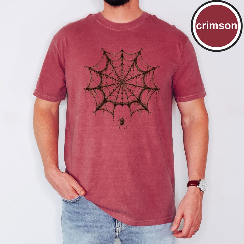 May include: A crimson short-sleeve t-shirt featuring a dark, detailed graphic of a spider web centered on the chest. A small spider is depicted hanging from the bottom of the web. The shirt has a crew neckline and a relaxed, casual fit.