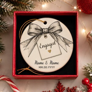 May include: A round white ceramic ornament featuring a black and white sketch of a bow tied to a tag with the text Engaged and a small gold heart. Below the tag, the text reads Name & Name and MM.DD.YYYY. The ornament has a gold hanging string.