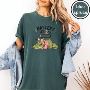 May include: A blue spruce t-shirt featuring a graphic of a tired green crocodile wearing a pink headscarf, with the text BATTERY AT 2% and a low battery icon above the crocodile. The shirt is worn with a light blue denim jacket over the shoulders.