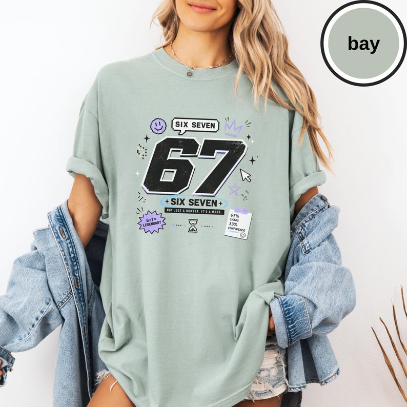 Pu&ograve; includere: Una t-shirt a maniche corte color verde salvia con una grande grafica del numero 67 in nero, il testo SIX SEVEN, 6+7=LEGENDARY e una piccola nota con scritto 67 CHAOS 33% CONFIDENCE. Il design include icone pixelate come uno smiley, una corona e una clessidra.