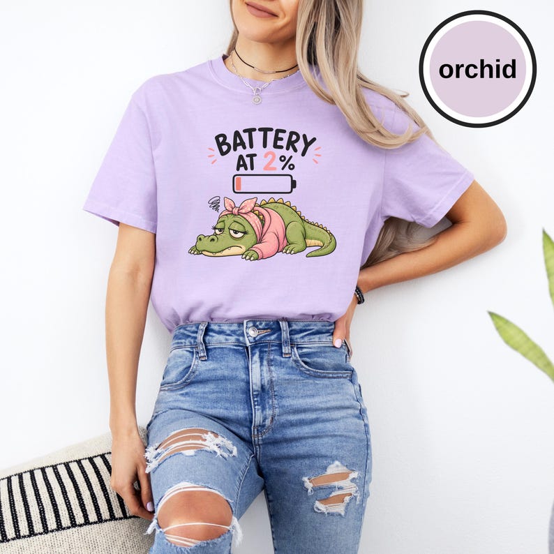 May include: A light purple t-shirt featuring a graphic of a tired green crocodile wearing a pink headband. Above the crocodile, the text reads BATTERY AT 2% with a low battery icon. The shirt has short sleeves and a crew neckline.